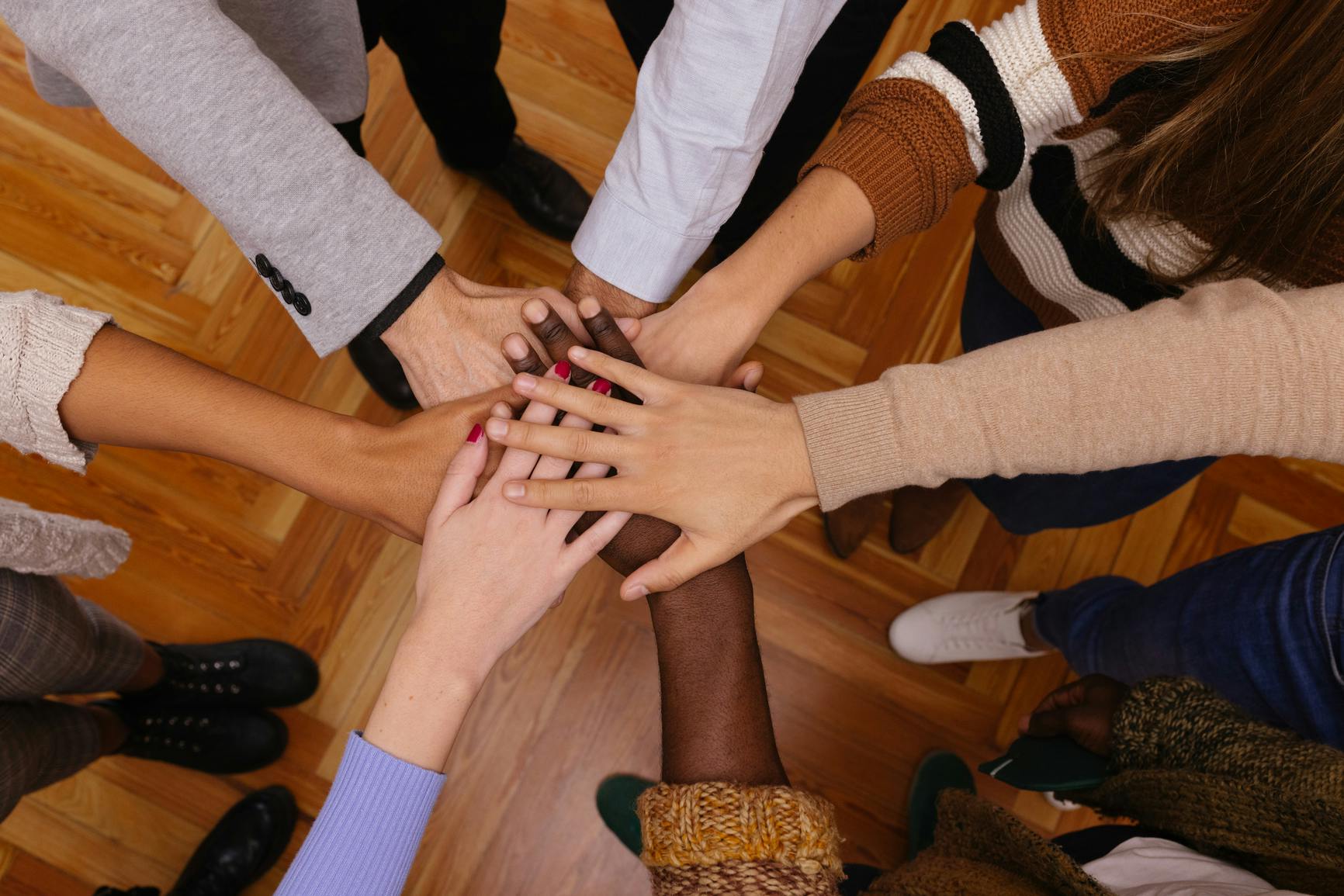 A closeup photograph of a group of people standing together with their arms pointing inward, creating a circle, their hands overlapping.