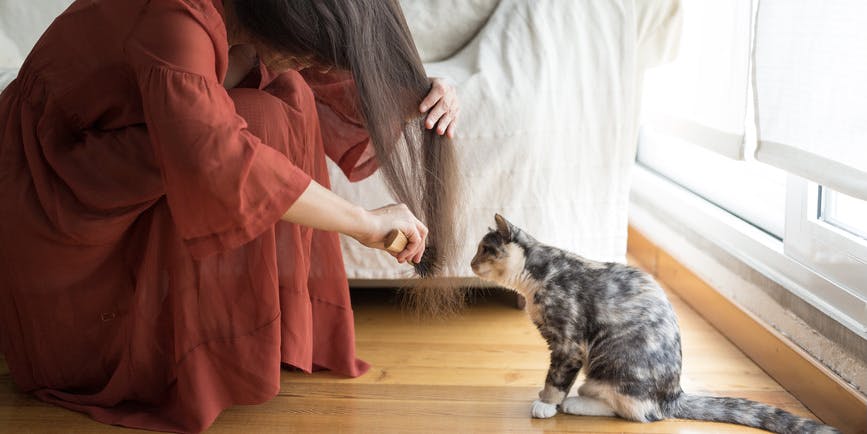 A woman wearing a long red dress squats low on the floor and brushes her long brown hair as a small multicolored cat looks at it curiously.