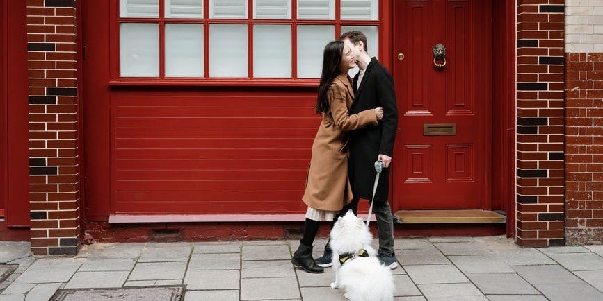 A color outdoor photograph of a sidewalk in front of a building with a bright red door and wall. A young Asian woman with long dark hair wearing a tan overcoat hugs a tall white man with brown hair wearing a black overcoat, their small white dog on a leash looking up at them.