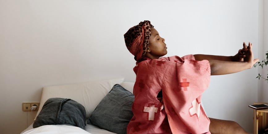 A color photograph of a Black woman in her bedroom, stretching while sitting up on her bed. She's wearing a pink-red nightdress and headband.