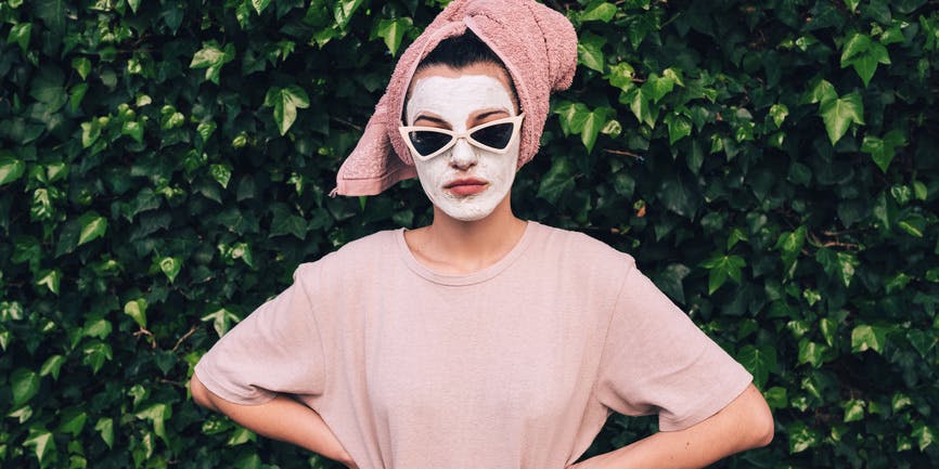 A young white woman with wearing a white skincare face mask, sunglasses, and a towel on her hair, stands against a wall of ivy and looks at the camera
