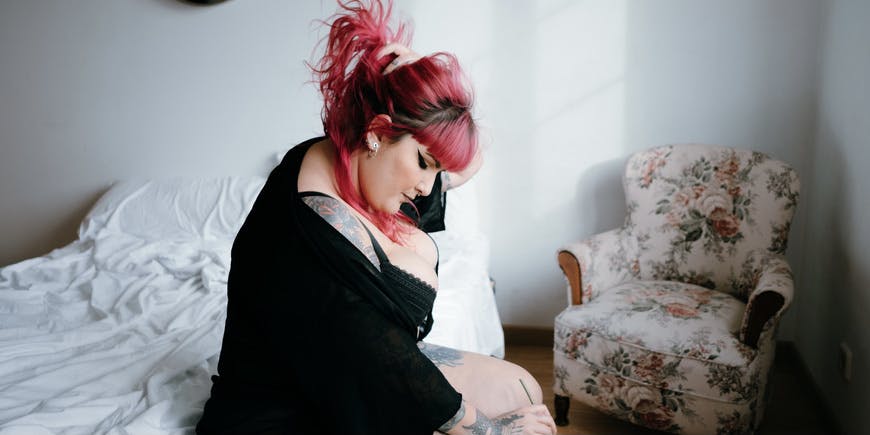 A curvy woman with pink hair wearing lingerie and a black sweater sits on her bed with her face tilted downward.