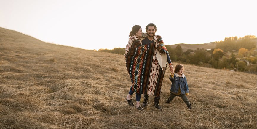 Family with little child covering themselves with geometric Ecuadorian blanket taking a walk by the countryside at sunset, spending quality time together in autumn