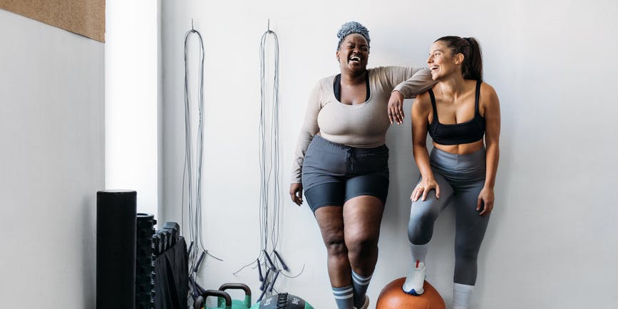 A curvy Black woman wearing gym clothes and a hair wrap and a muscular white woman wearing gym clothes stand close together, laughing, resting against a well of a gym.