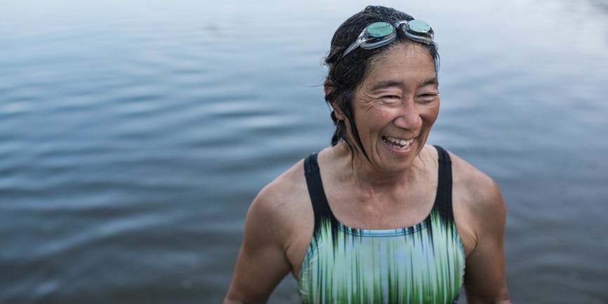 Portrait of a tanned woman in her fifties or sixties wearing a swimsuit and goggles, smiling as she comes out of swimming in a lake.