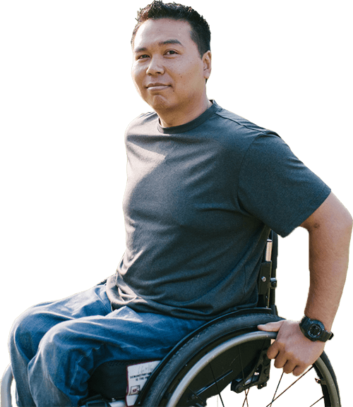 Asian man in wheel chair, sitting in sun on grass, hands on wheels, smiling subtly at camera.