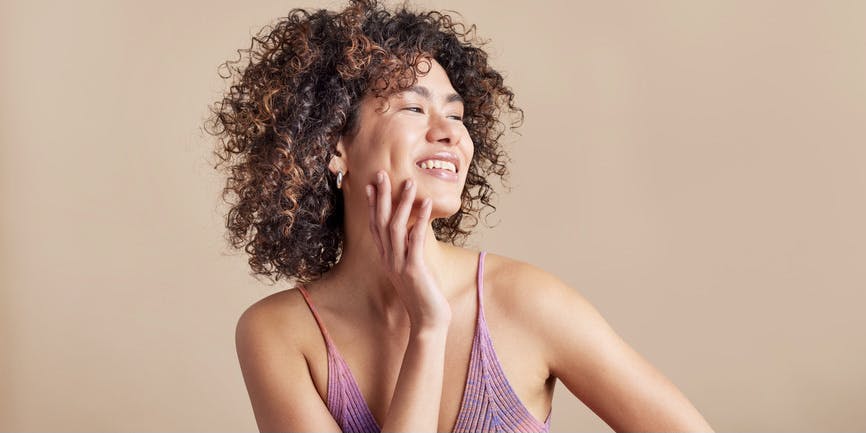 A young woman with brown skin and brown Afro hair smiles while touching her cheek and looking off camera. She’s wearing a purple tank and standing against a neutral colored wall.
