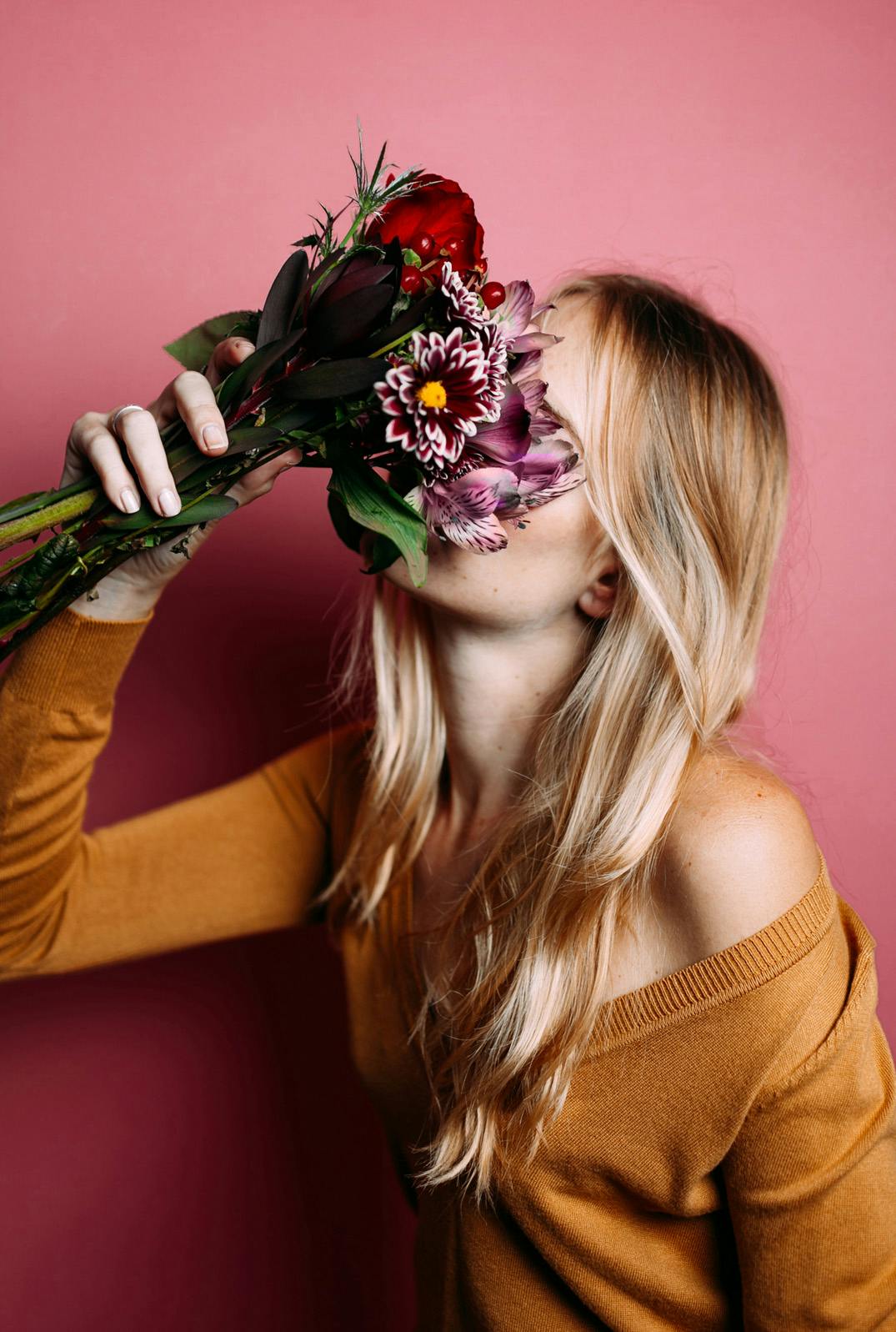 A woman with blonde burying her face into a bouquet flowers, standing against a red wall