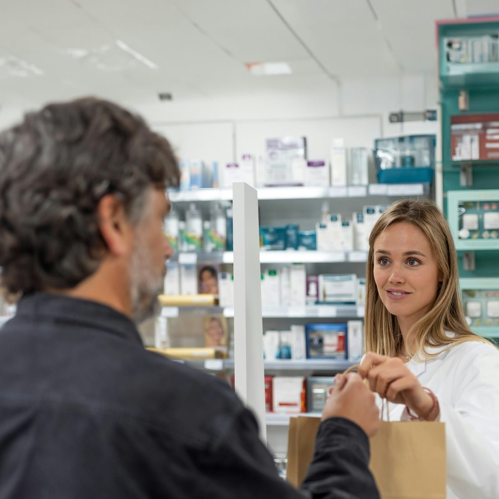A graying man picking up a prescription from a smiling pharmacist, who is handing a bag to him over the pharmacy counter.