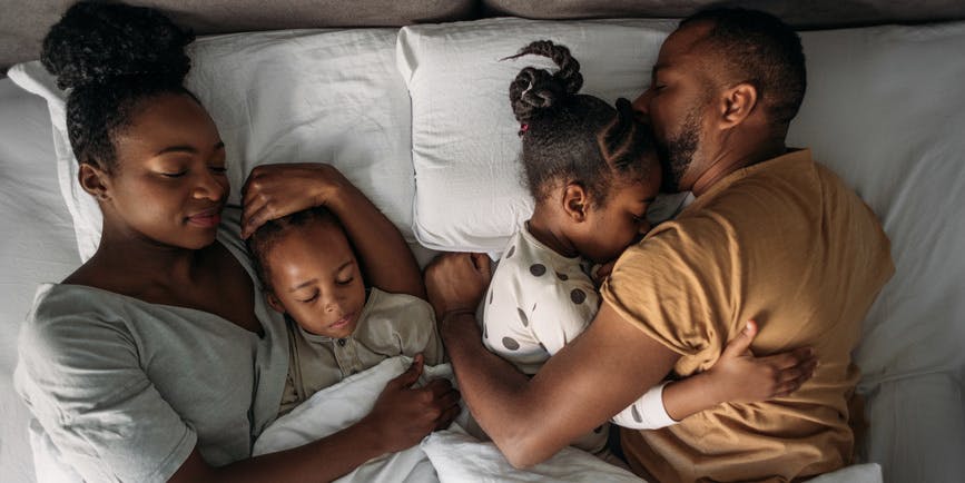 Photograph from above of a Black family in bed, with a man and woman cuddling two small children.