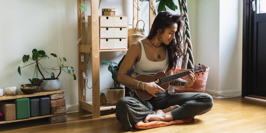 Relaxed young woman with brown skin and long brown hair enjoys playing a ukulele while sitting on the floor of her clean, airy home.